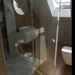 Nowoczesna łazienka na poddaszu z kabiną prysznicową, umywalką nablatową, okrągłym lustrem i toaletą, wykończona płytkami imitującymi marmur i drewno, oświetlona światłem dziennym z okna dachowego.