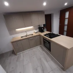 Jasna kuchnia z drewnianymi frontami, wyposażona w płytę gazową, piekarnik i ekspres do kawy, z widoczną osobą w tle.