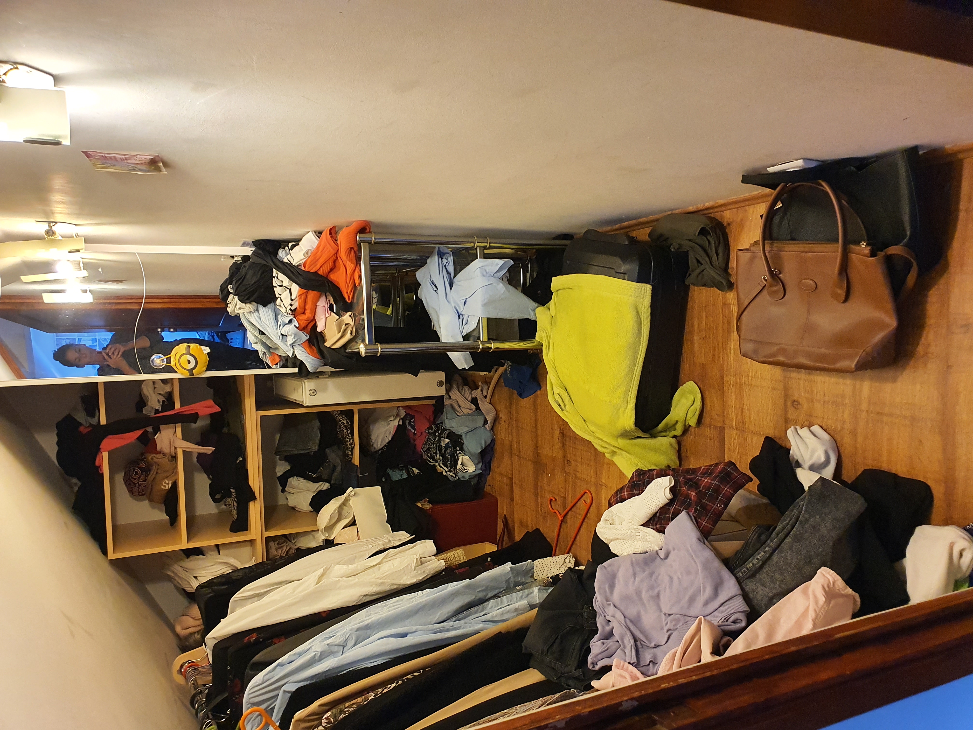 Przepełniona garderoba z ubraniami na półkach, w szafie i na podłodze, z torebkami i walizką na górze.