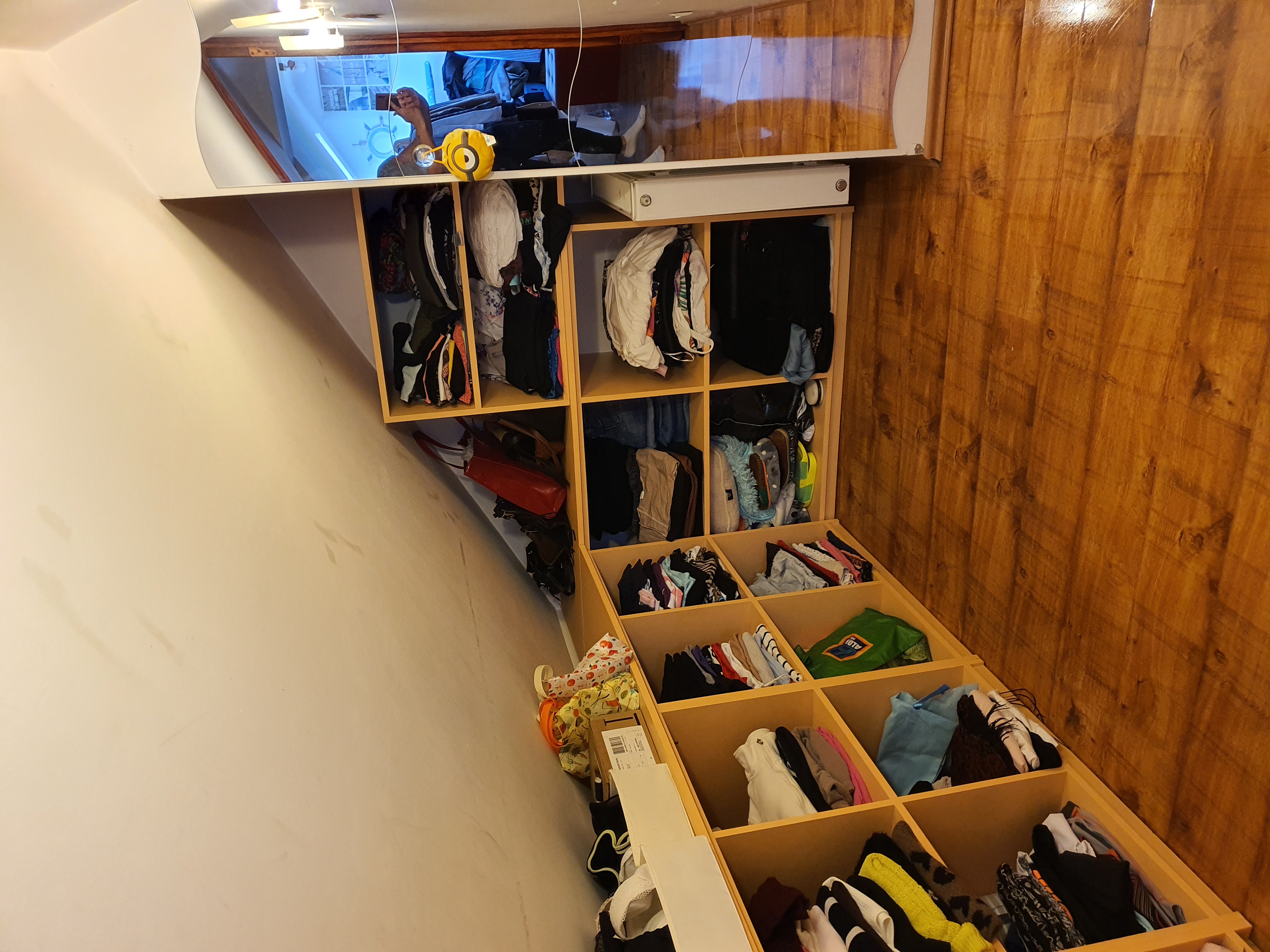 Uporządkowana garderoba z półkami wypełnionymi ubraniami, torbami i akcesoriami, oświetlona od góry, z widocznym odbiciem osoby robiącej zdjęcie w przeszklonej szafce.