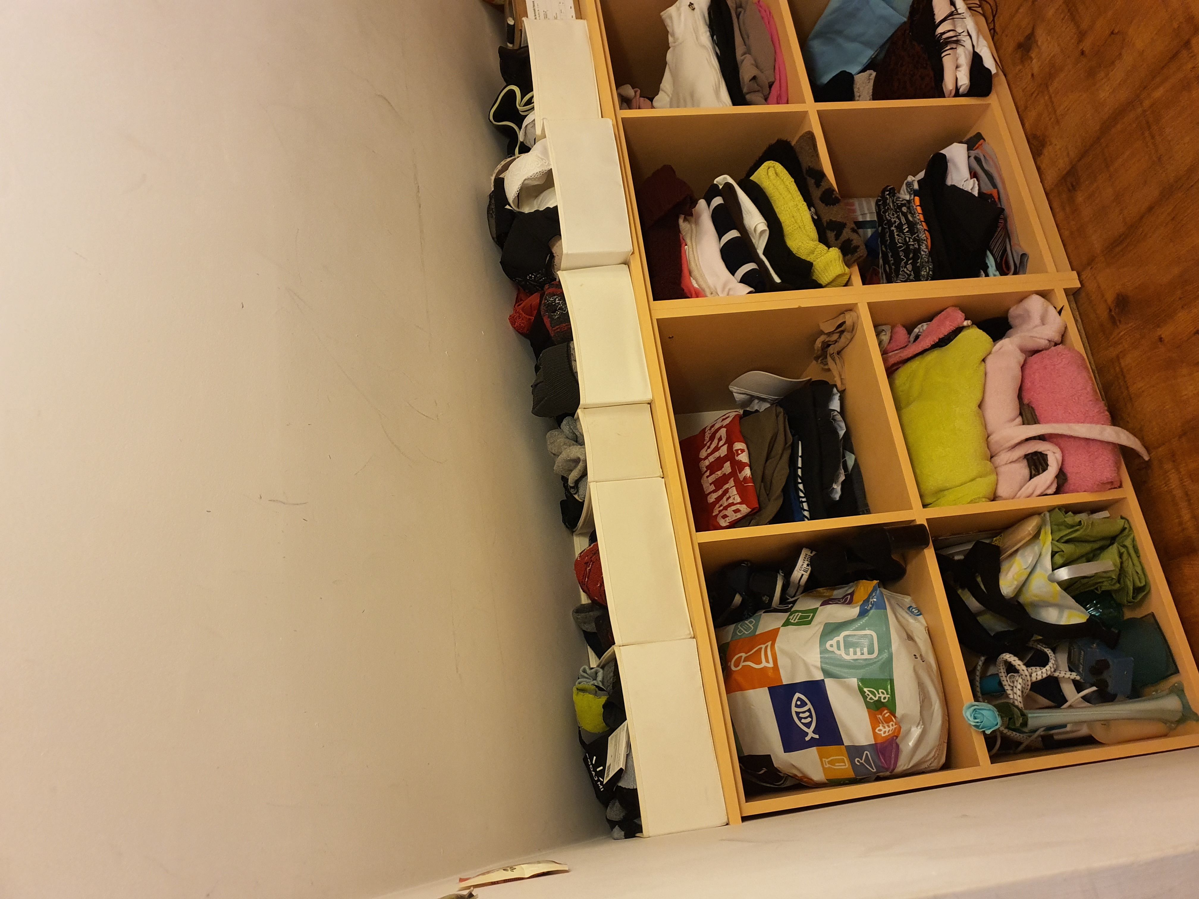 Wnętrze szafy z półkami wypełnionymi ubraniami, tekstyliami i torbą na zakupy z kolorowymi wzorami, widok z dołu.