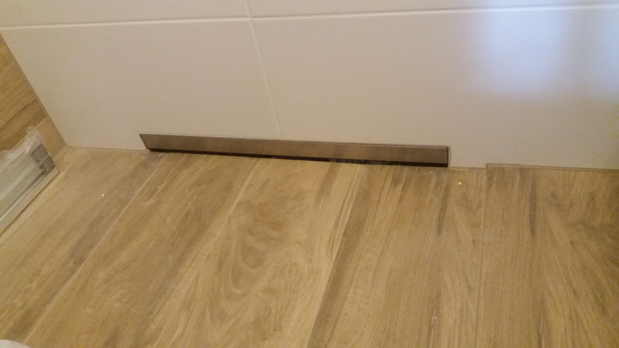 Szczegółowe ujęcie podłogi z paneli drewnopodobnych i białych płytek ściennych z metalową listwą przypodłogową; widoczne nierówności w ułożeniu paneli.