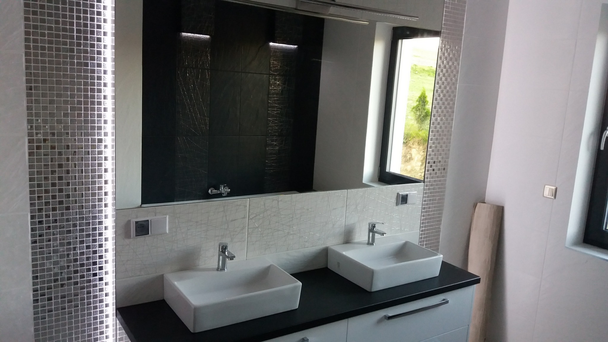 Nowoczesna łazienka z dwoma prostokątnymi umywalkami nablatowymi, czarnym blatem, lustrem na całą szerokość ściany, podświetlaną mozaiką i czarnymi płytkami w strefie prysznica.