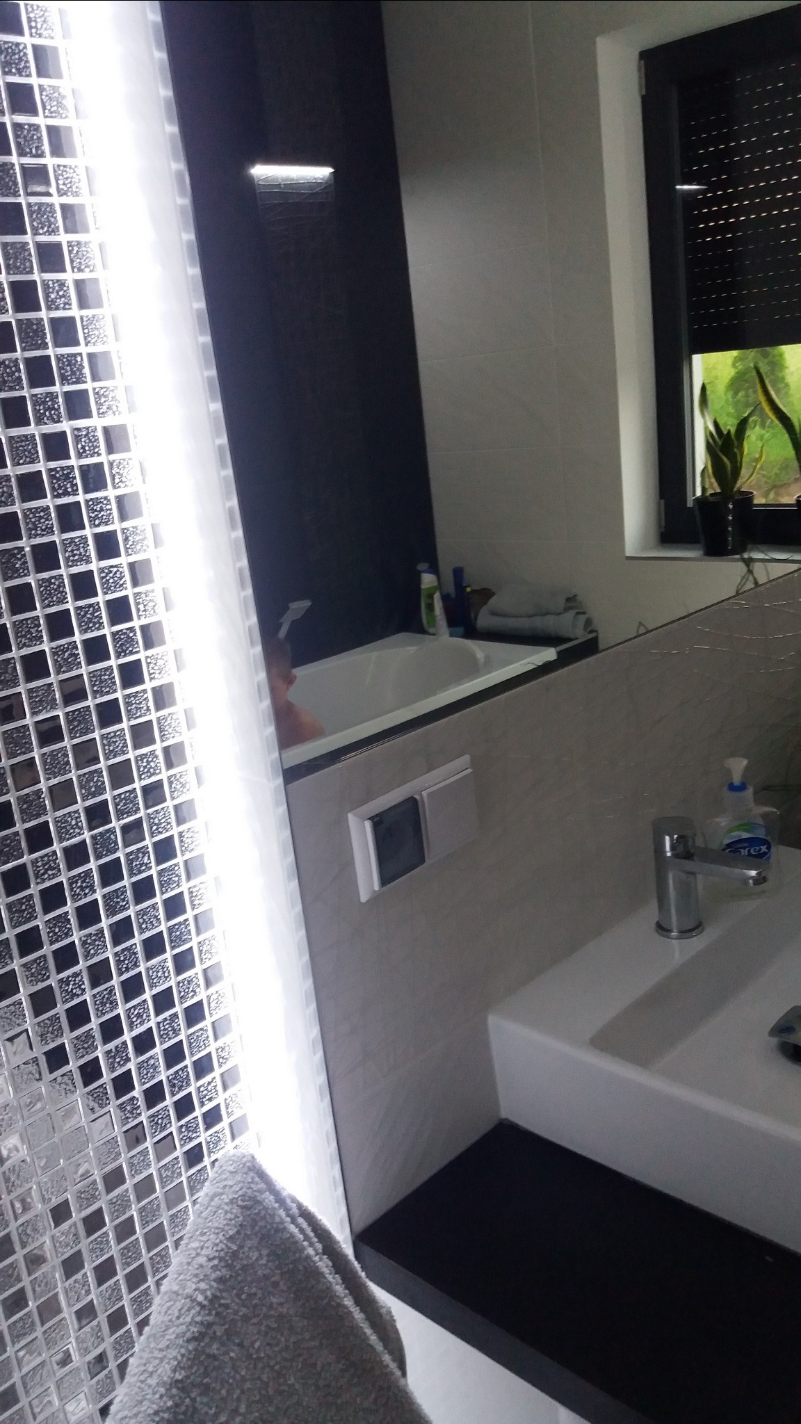 Nowoczesna łazienka z mozaiką w odcieniach szarości, białą umywalką z czarnym blatem, baterią i oknem z roletami w tle.