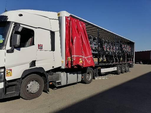 Biała ciężarówka z czerwoną plandeką, załadowana dużą ilością czarnych maszyn rolniczych, stojąca na betonowym placu w słoneczny dzień.