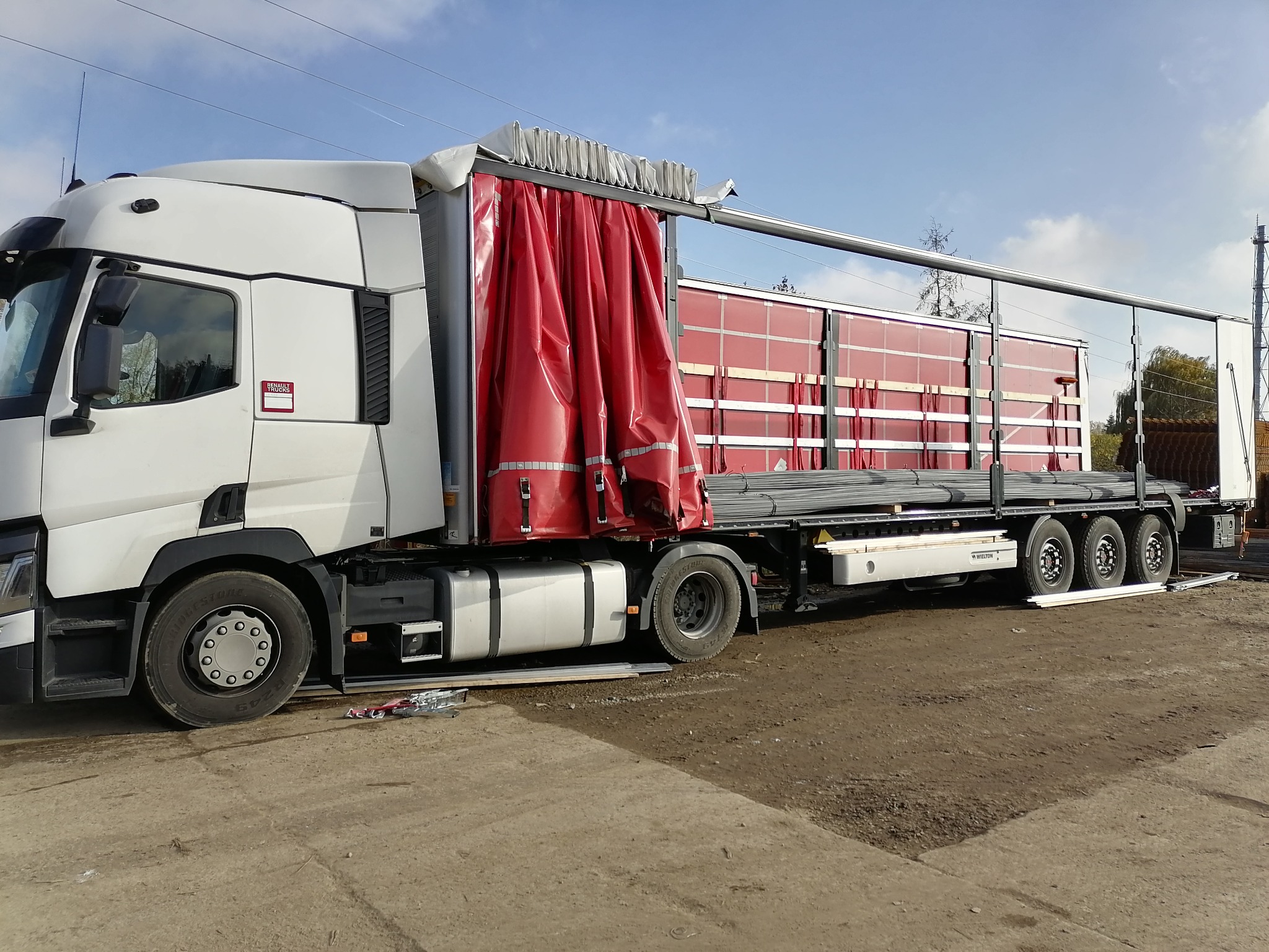 Ciężarówka Renault z czerwoną plandeką, załadowana stalowymi prętami, widoczna na placu budowy w słoneczny dzień.