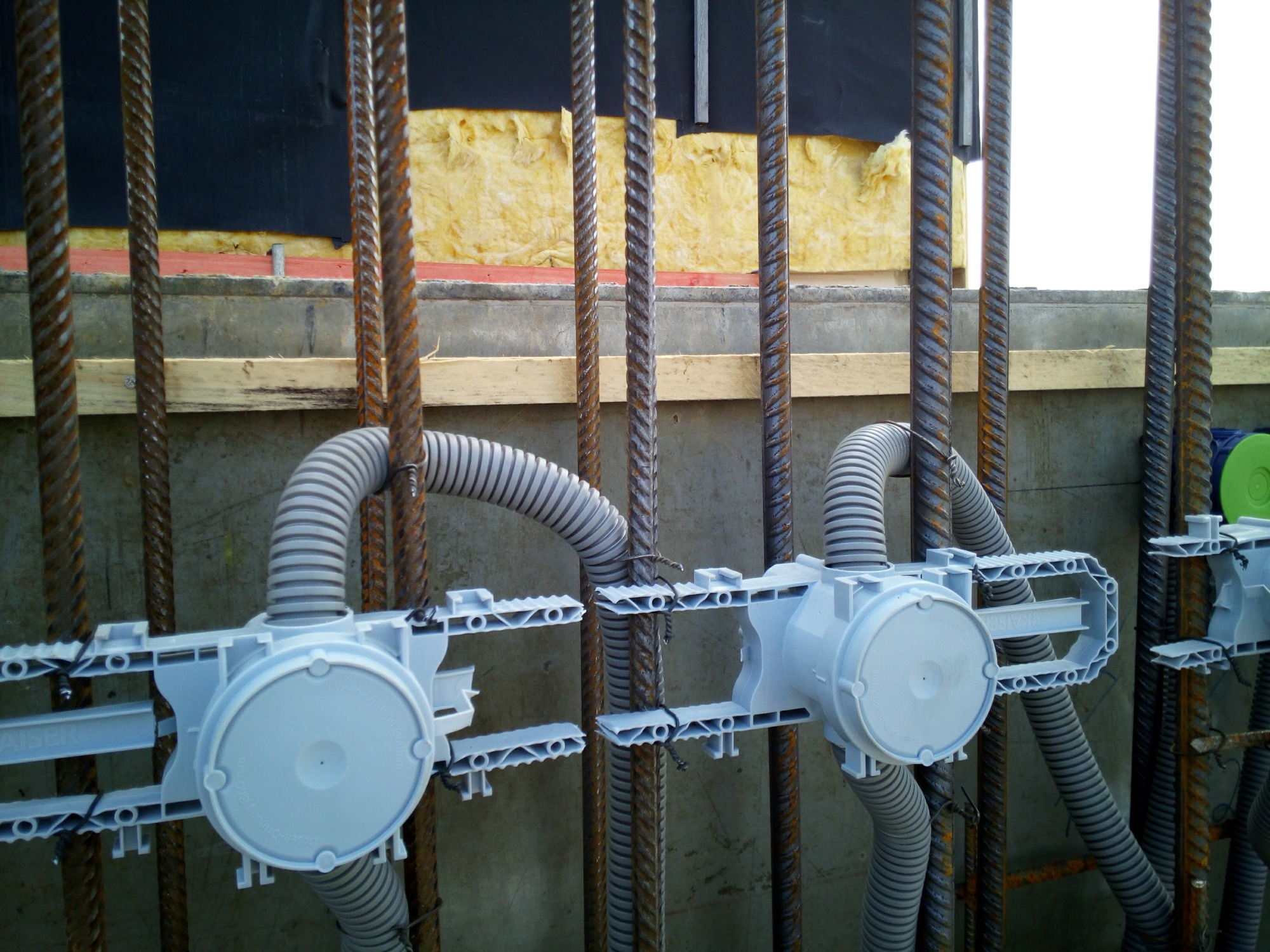 Szare rury karbowane i puszki instalacyjne przymocowane drutem do żelbetowej konstrukcji z izolacją z wełny mineralnej w tle.