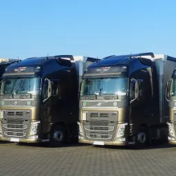 Cztery ciężarówki Volvo w kolorze brązowo-czarnym, zaparkowane równo na szarym bruku, z logo firmy 'Tra-Met' widocznym na przedniej szybie, w słoneczny dzień.