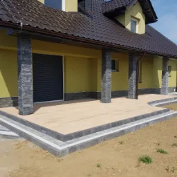 Żółty dom z brązowym dachem i tarasem wyłożonym jasnymi płytami, obrzeża z szarej kostki, słupy z kamienia, rolety zewnętrzne.