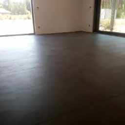 Ułożona, ciemnoszara podłoga z płytek wielkoformatowych w surowym wnętrzu z dużymi oknami i widokiem na zewnątrz, widoczne ślady po remoncie.