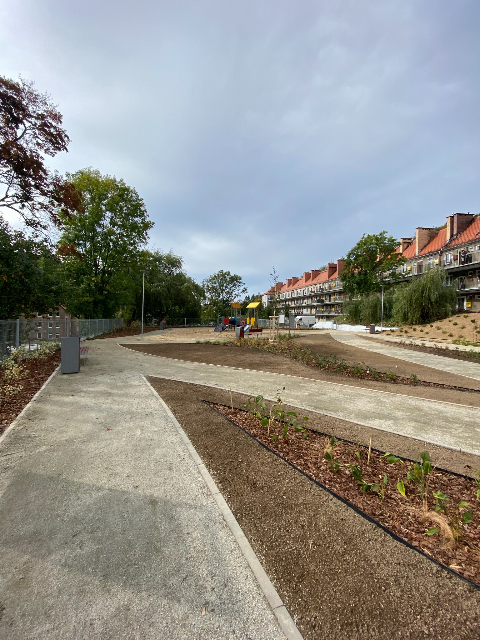 Nowo założony park kieszonkowy: betonowe ścieżki, rabaty z korą i młodymi roślinami, plac zabaw w tle, budynki mieszkalne w Wałbrzychu.