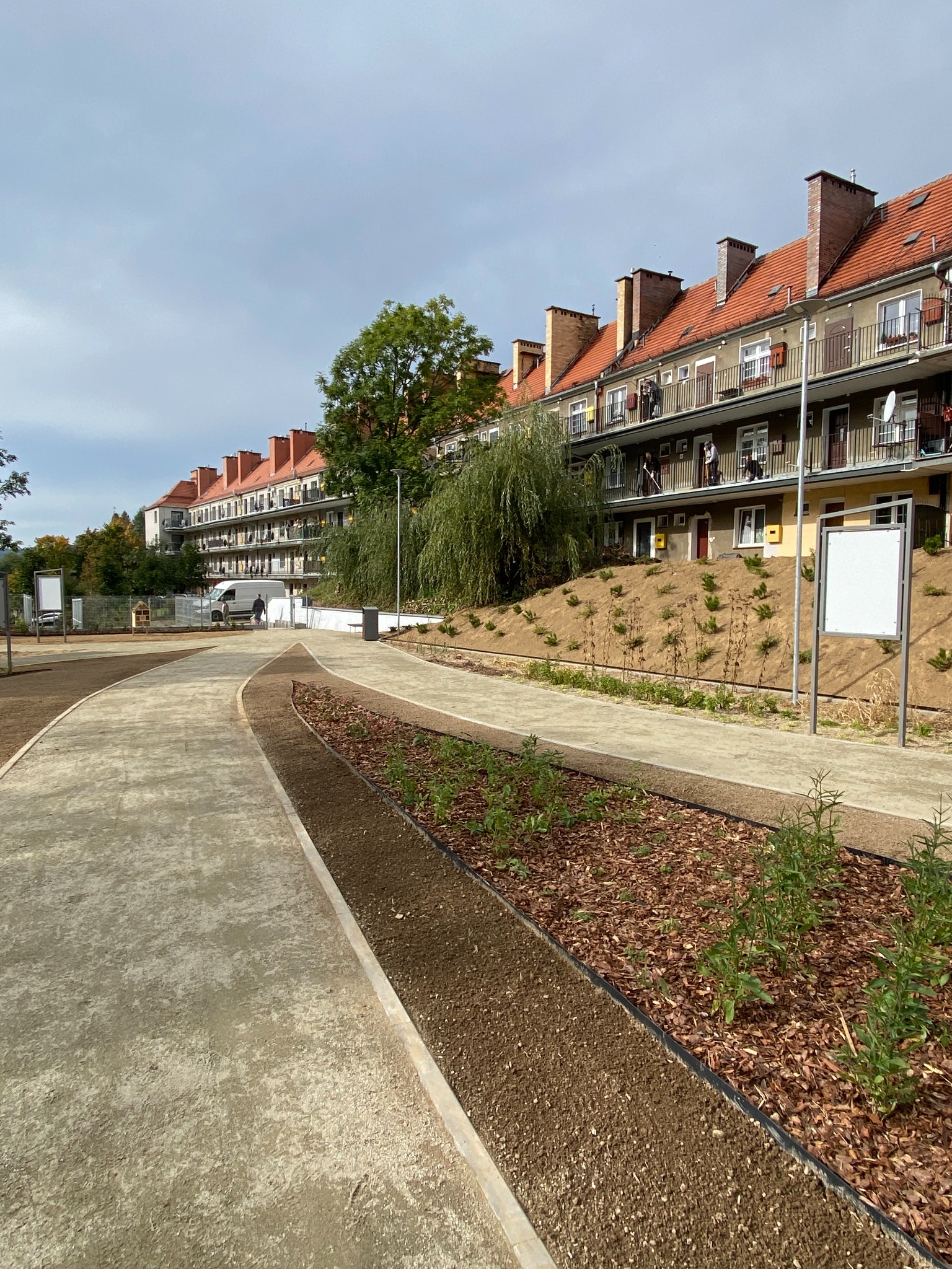 Nowo zagospodarowany teren zielony w miejskiej przestrzeni, z wytyczonymi ścieżkami, rabatami z korą i młodymi roślinami, na tle bloku mieszkalnego z balkonami i czerwonym dachem, Wałbrzych.