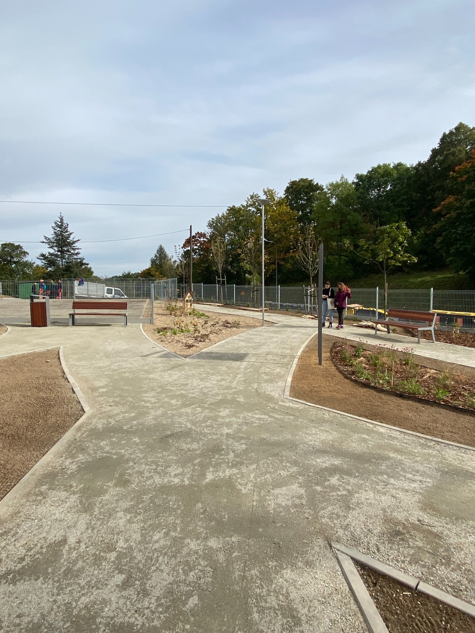 Nowo wybudowany park kieszonkowy z betonowymi ścieżkami, drewnianymi ławkami, rabatami z młodymi roślinami i metalowym ogrodzeniem, widok na drzewa w tle.
