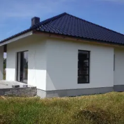 Nowo otynkowany, parterowy dom z ciemną dachówką i czarnymi ramami okiennymi, widok z zewnątrz na częściowo wykończony taras z bloczków betonowych i zieloną rośliną doniczkową.