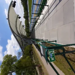 Zadaszone trybuny stadionu z zielonymi barierkami i płytami chodnikowymi, znak ewakuacyjny z prawej strony, w tle bieżnia i drzewa.