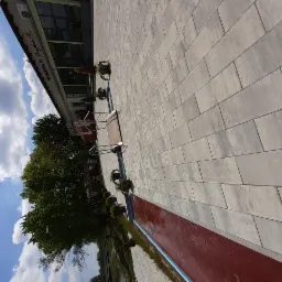 Nowoczesna nawierzchnia z jasnoszarej kostki brukowej przed budynkiem restauracji z kwiatami w donicach, widok z góry na fragment chodnika i czerwoną obudowę rabaty.