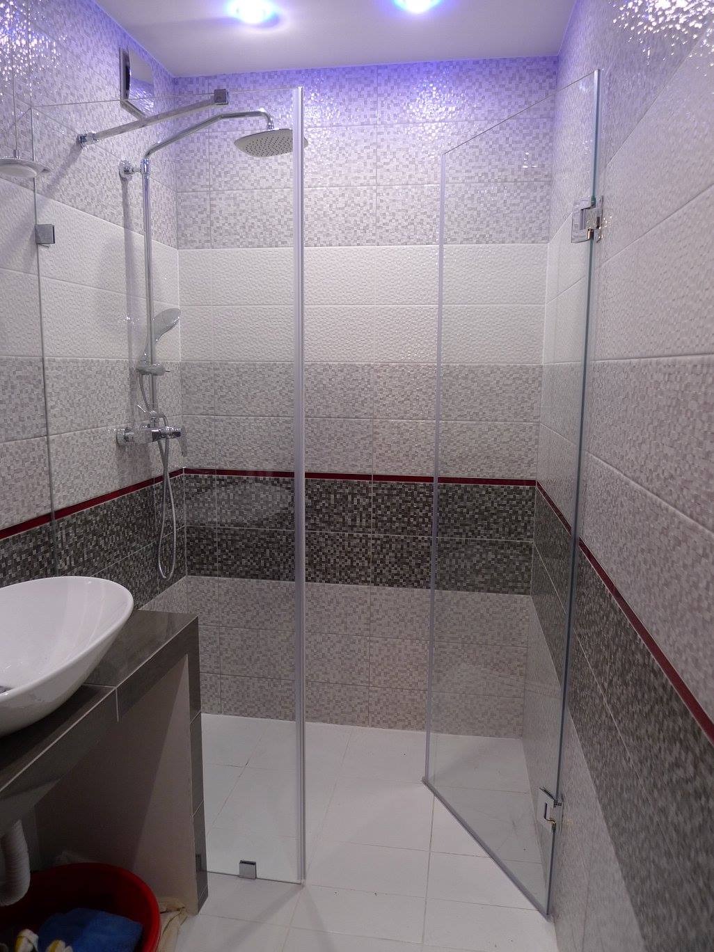 Nowoczesna kabina prysznicowa z przezroczystego szkła, z widocznymi chromowanymi zawiasami i deszczownicą, na tle mozaikowej ściany w odcieniach szarości i bieli.