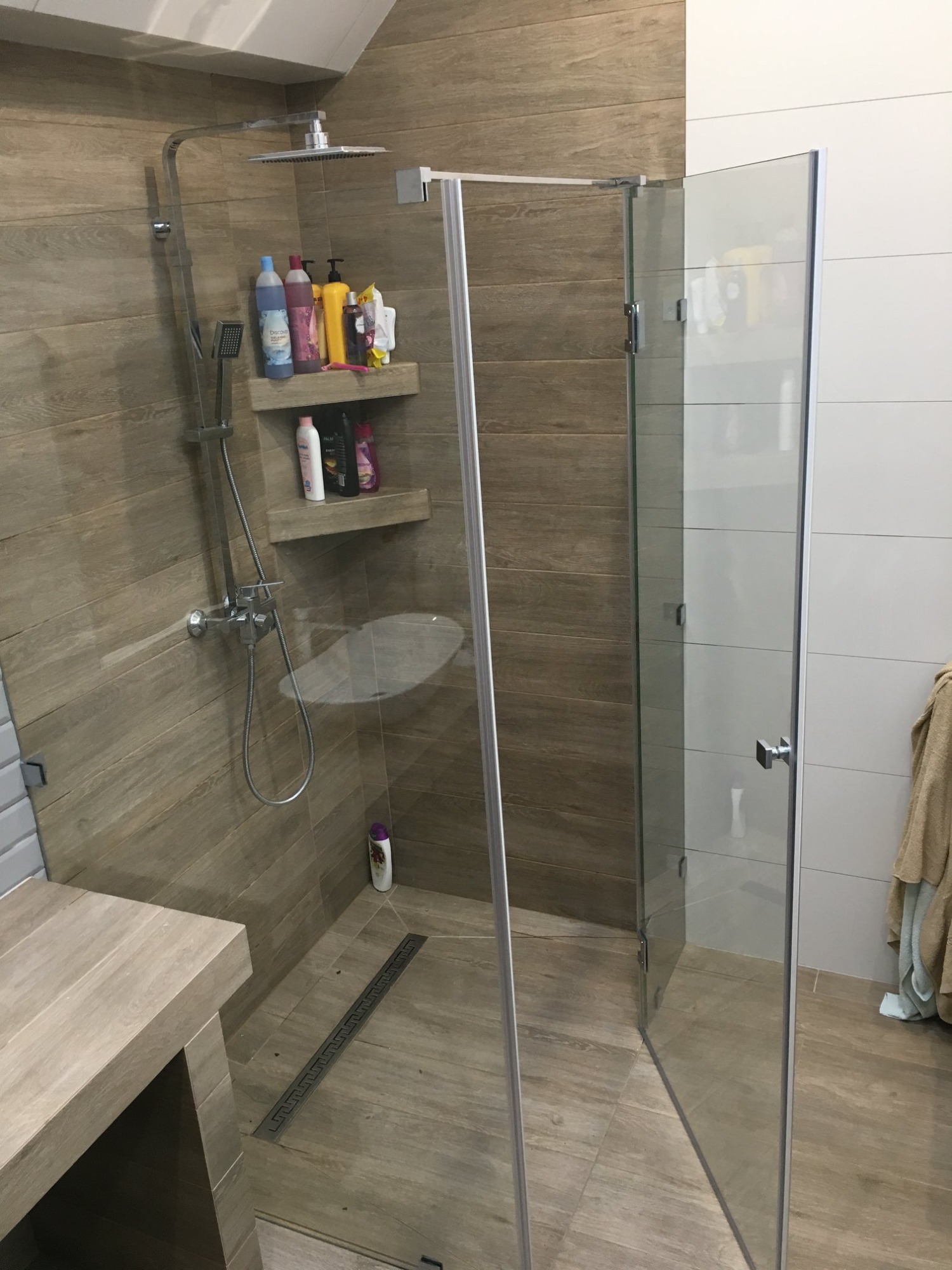 Otwarta kabina prysznicowa z szklanymi drzwiami, wyłożona płytkami imitującymi drewno, z półkami na kosmetyki i chromowaną armaturą.