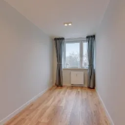 Pusty pokój po malowaniu, z drewnopodobną podłogą, szarymi zasłonami i białym grzejnikiem pod oknem.