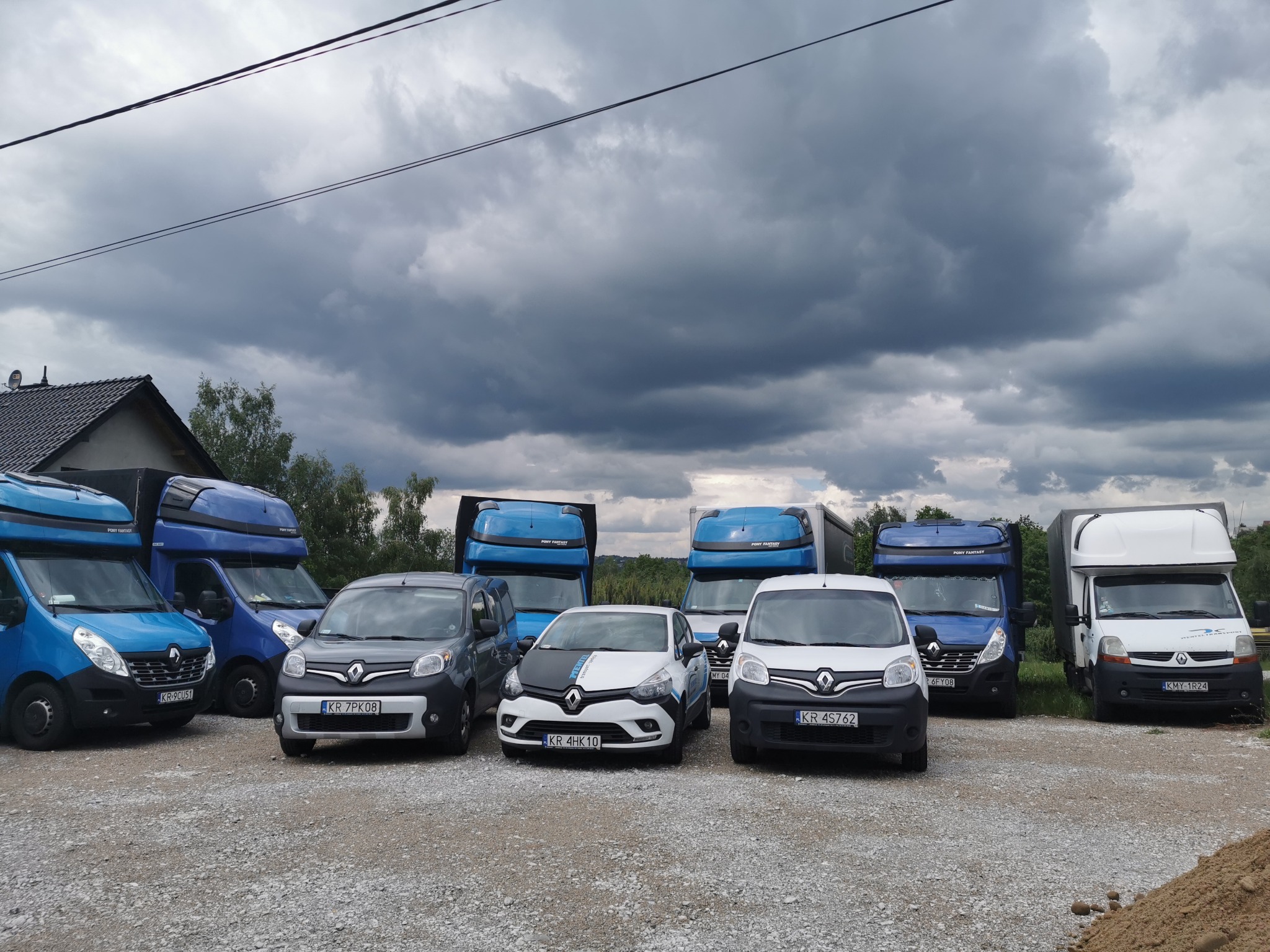 Flota samochodów transportowych różnych rozmiarów, od osobowych po ciężarowe, zaparkowana na żwirowym placu pod pochmurnym niebem.