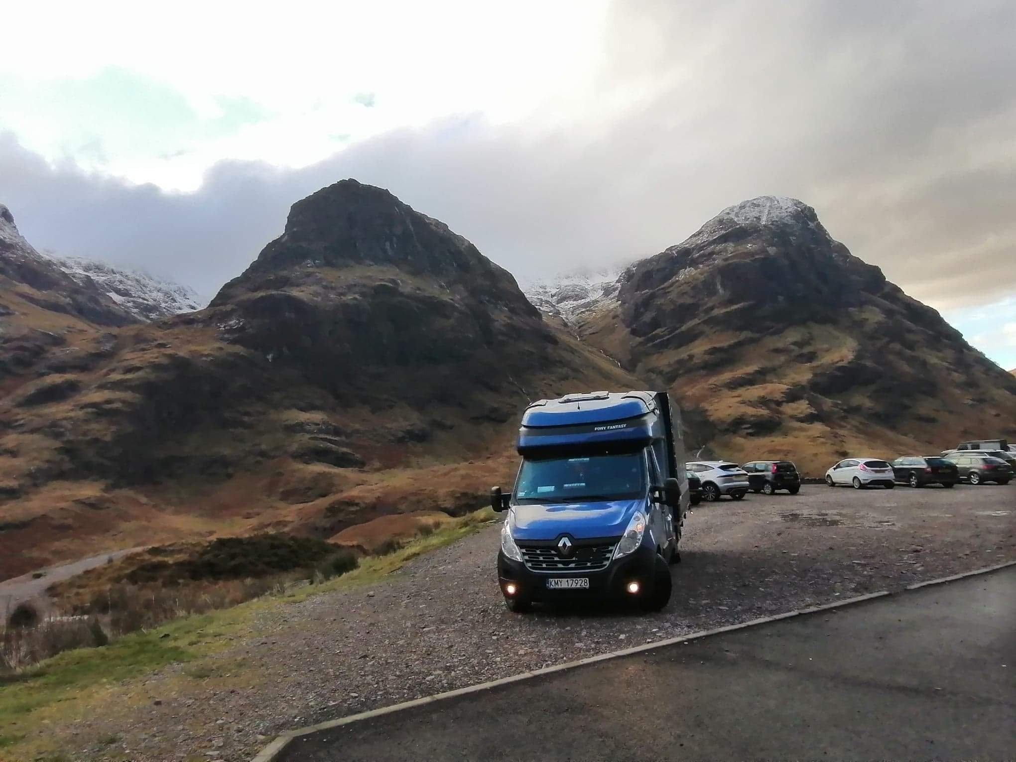 Niebieski bus transportowy Renault Master z nadbudową Pony Fantasy parkuje na żwirowym parkingu z widokiem na góry z ośnieżonymi szczytami w pochmurny dzień.