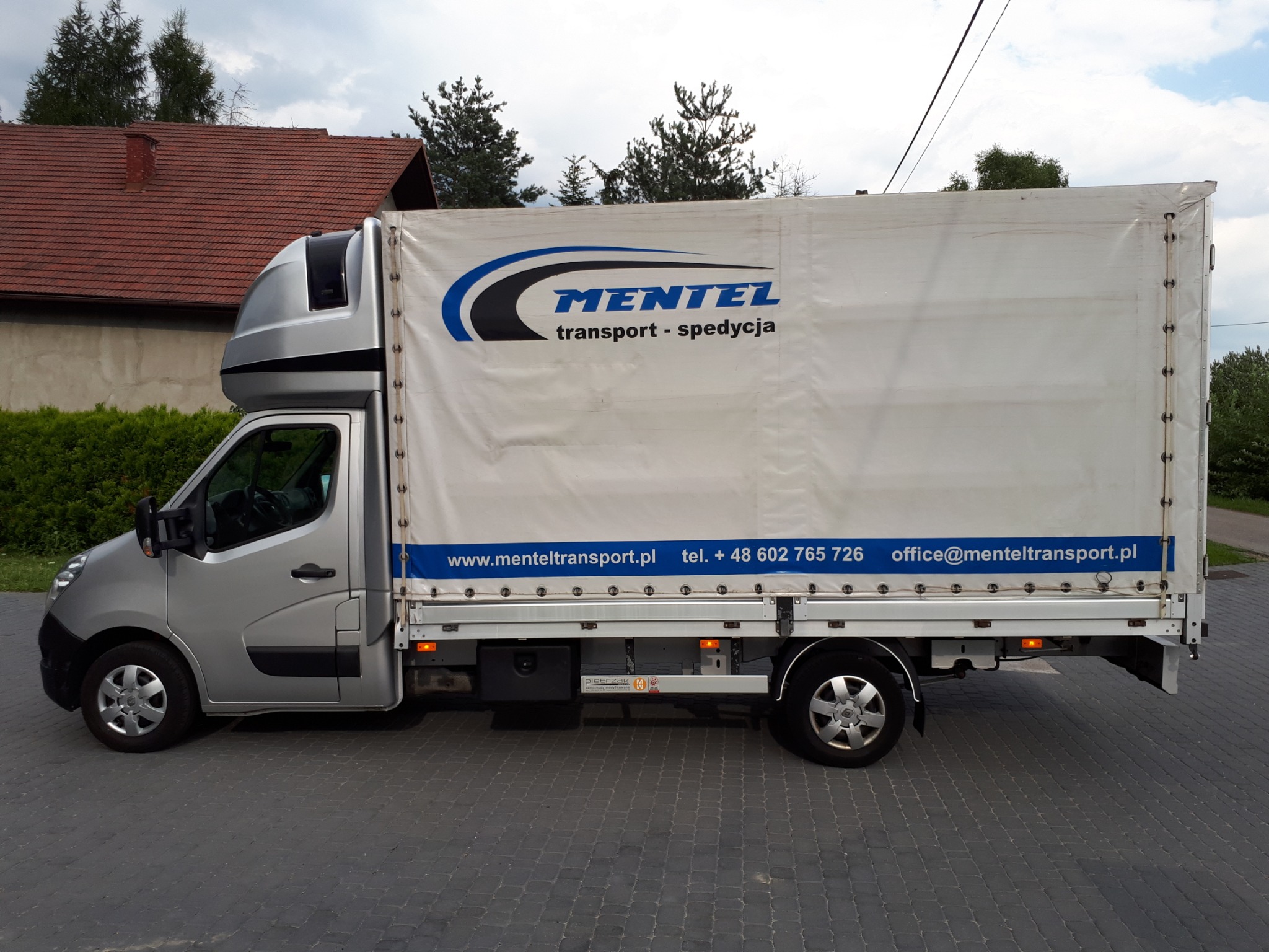 Srebrny samochód dostawczy z plandeką firmy Mentel Transport zaparkowany na brukowanej ulicy, widoczne logo i dane kontaktowe na plandece.