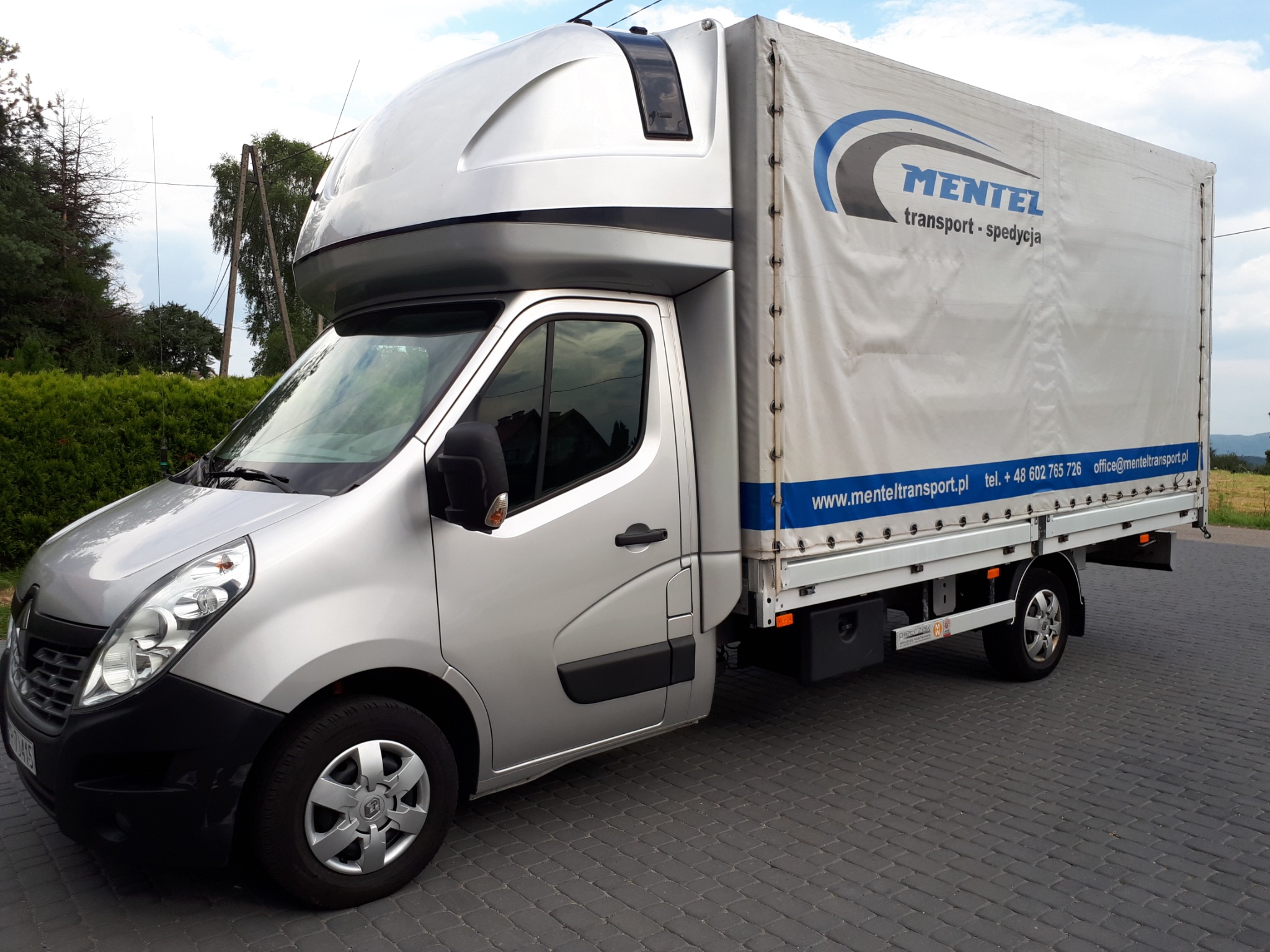 Srebrny samochód dostawczy Renault Master z plandeką firmy Mentel Transport, stojący na kostce brukowej, widok z boku.