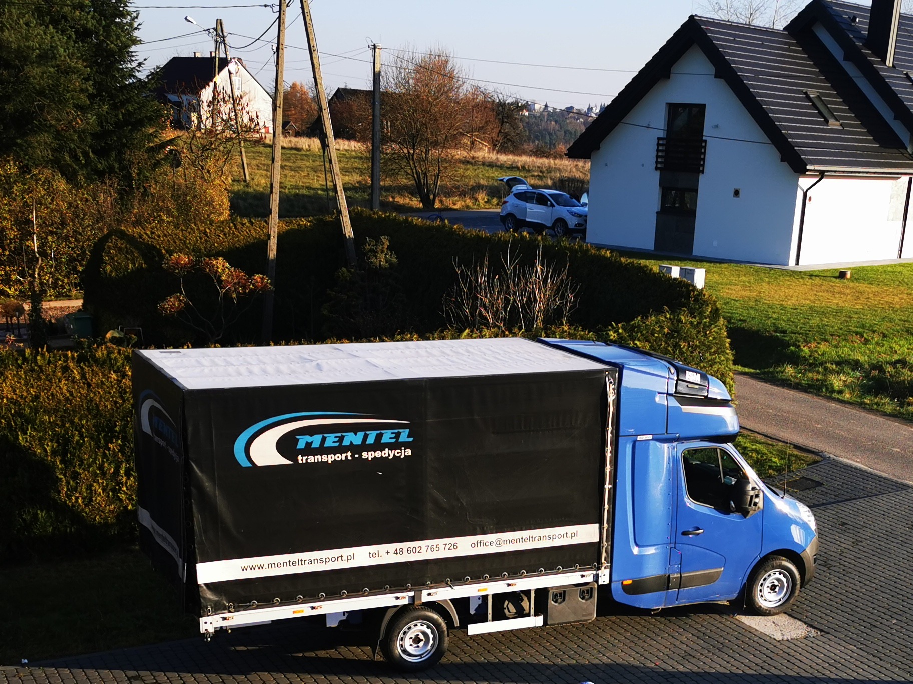 Niebieska ciężarówka z plandeką firmy transportowej z Sieprawia, z logo i danymi kontaktowymi, zaparkowana na brukowanej drodze przed nowoczesnym domem w słoneczny dzień.