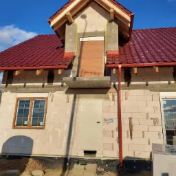 Dom w trakcie budowy z zamontowanymi oknami z brązowymi ramami, częściowo zamontowanymi roletami zewnętrznymi i czerwonym dachem z rynnami.