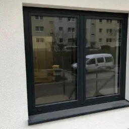 Nowe, grafitowe okno dwuskrzydłowe z widokiem na budynek mieszkalny i zaparkowany biały samochód dostawczy.