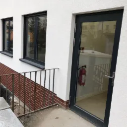 Budynek z białym tynkiem, ciemne okna i drzwi wejściowe z widocznym wewnątrz budynku sprzętem przeciwpożarowym, fragment ceglanej podmurówki i metalowej balustrady.