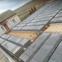 Wieniec stropowy z szalunkiem traconym, zbrojony siatką stalową, przygotowany do zalania betonem, widok z góry na budowę domu jednorodzinnego na wsi, pochmurne niebo w tle.