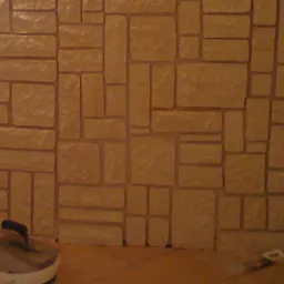 Ściana wykończona imitacją kamienia w odcieniach beżu, z widocznym wiaderkiem i kielnią u dołu.