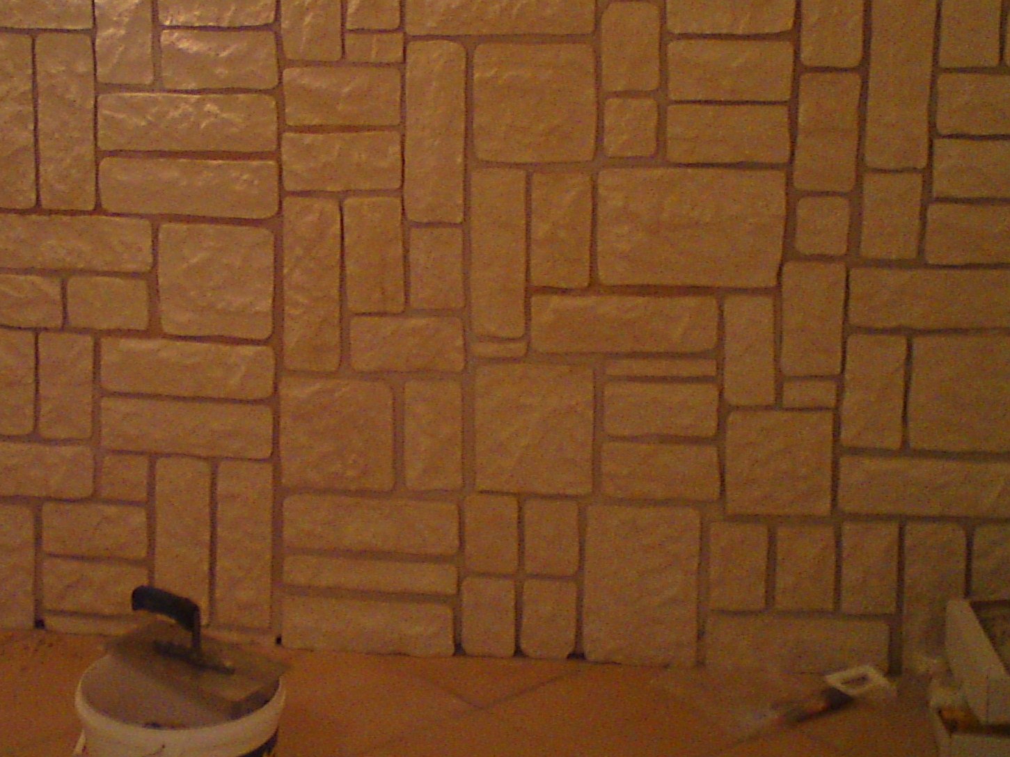 Ściana wykończona imitacją kamienia w odcieniach beżu, z widocznym wiaderkiem i kielnią u dołu.