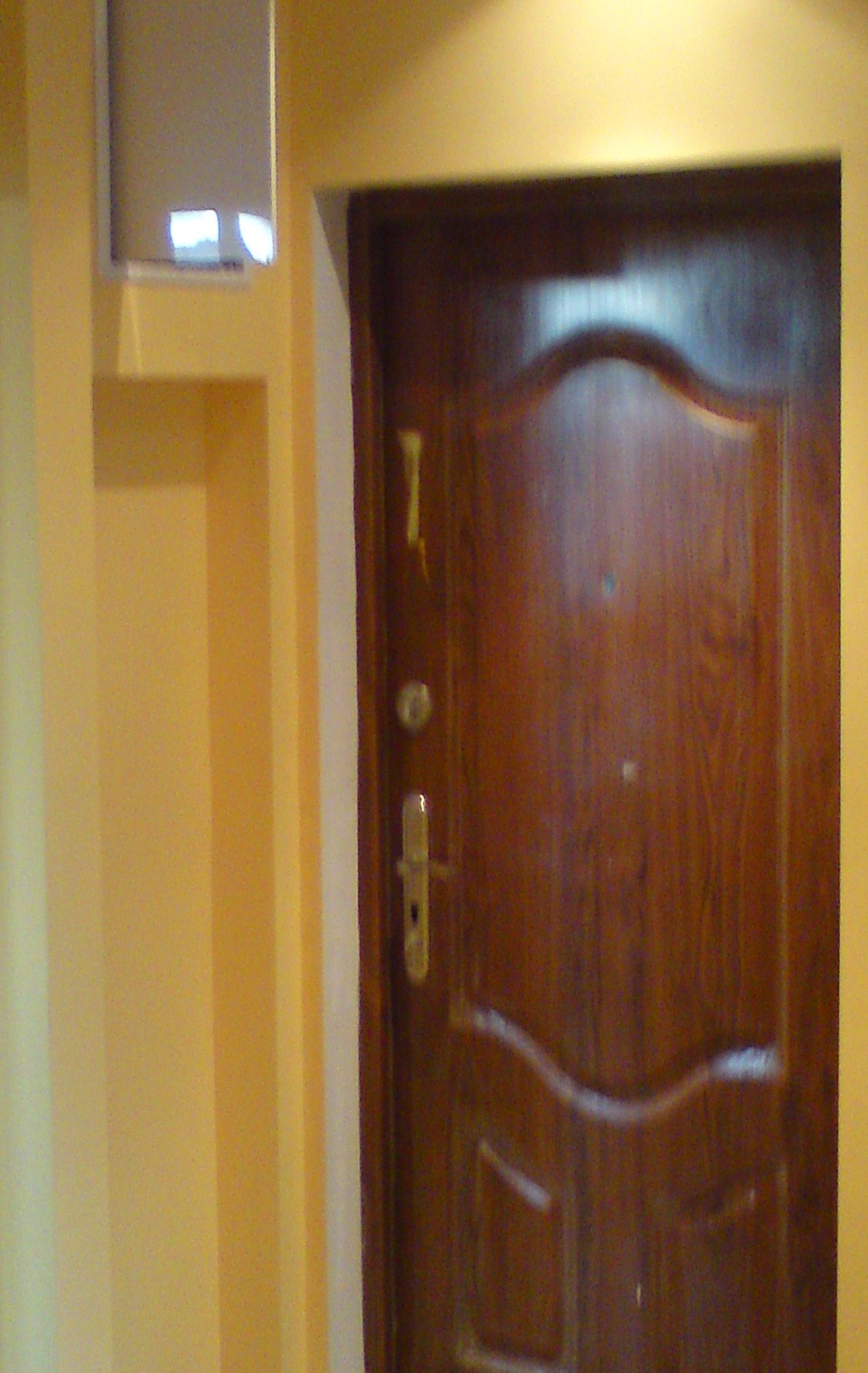 Ciemnobrązowe drzwi wejściowe z wizjerem i złotą klamką, osadzone w ościeżnicy w pomarańczowo-żółtej ścianie z widocznym fragmentem rolety okiennej w górnym lewym rogu.