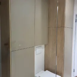 Nowoczesna łazienka z zabudową meblową w odcieniach beżu i drewna, ukrywająca spłuczkę toalety, z minimalistycznym designem i białym sanitariatem.