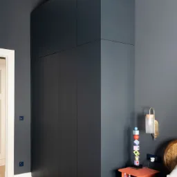 Wysoka, zabudowana szafa wnękowa w kolorze grafitowym, sięgająca sufitu, w sypialni z parkietem w jodełkę i ciemnymi ścianami, obok łóżka z rattanowym zagłówkiem i pomarańczową szafką nocną z lampką.