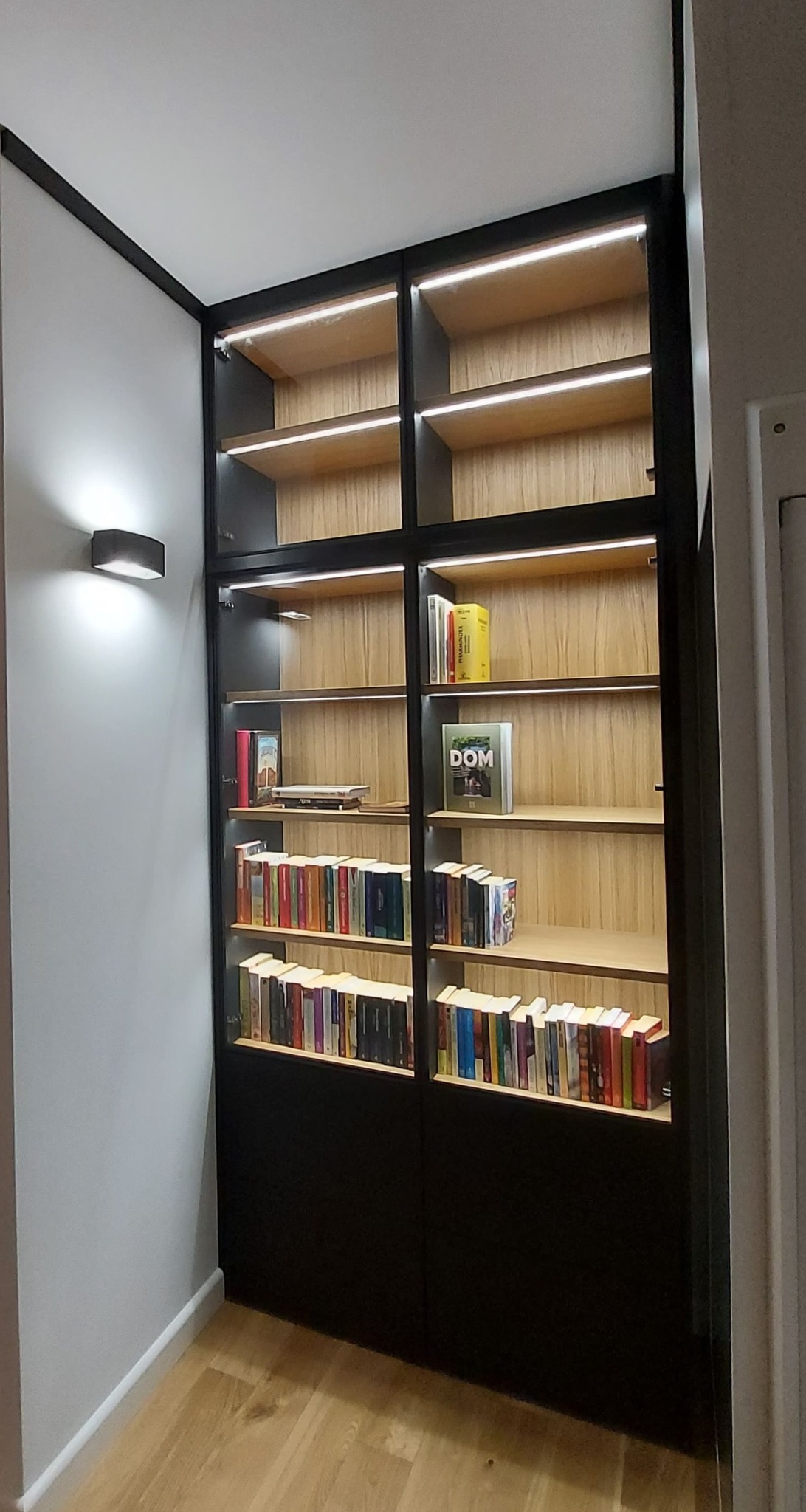 Wysoka, wbudowana szafa z drewnianymi półkami, oświetleniem LED i czarną ramą, wypełniona książkami, umieszczona we wnęce w pokoju z parkietem.