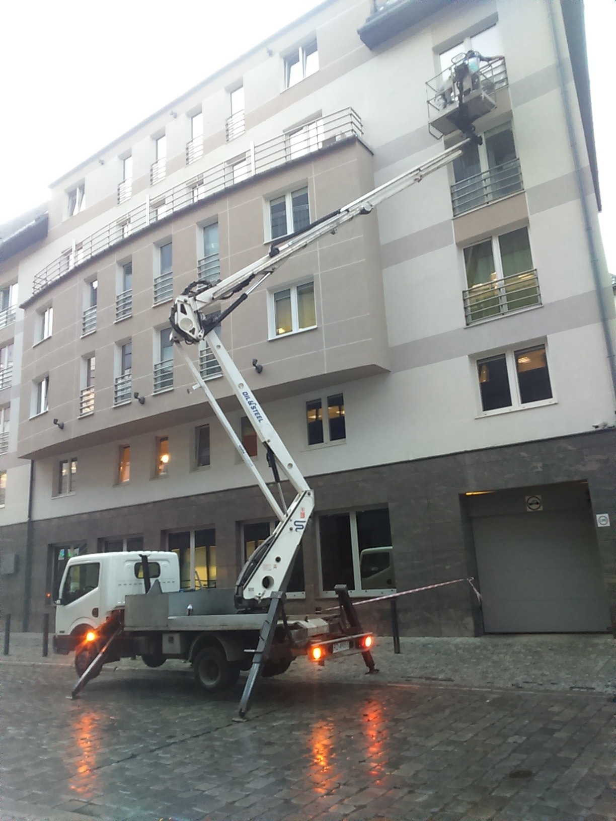 Pracownik w koszu podnośnika teleskopowego, wykonujący prace na elewacji budynku mieszkalnego z balkonami, widok na mokry chodnik z kostki brukowej.