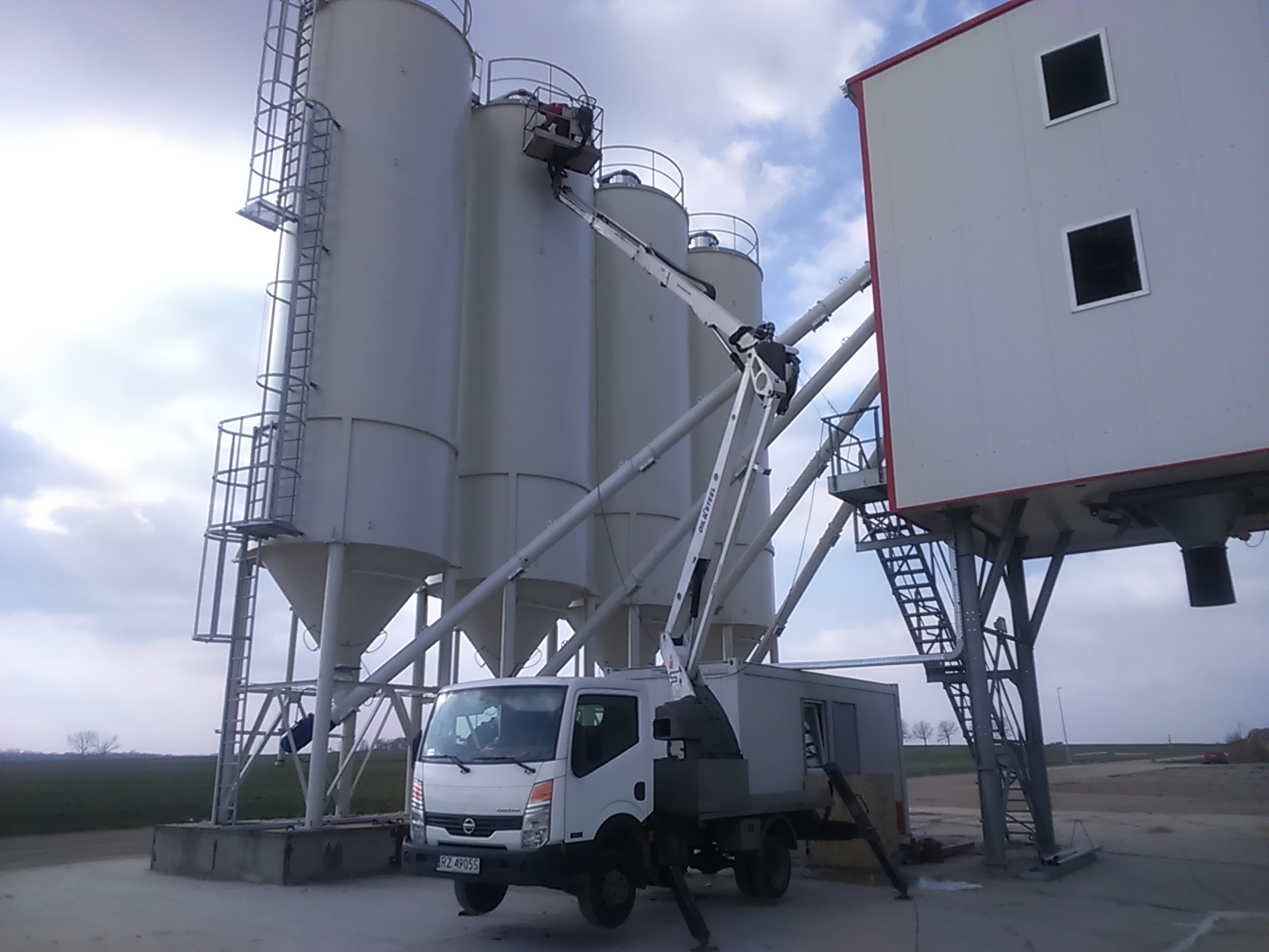 Biała ciężarówka z podnośnikiem koszowym przy silosach i konstrukcji wsporczej, prawdopodobnie podczas prac konserwacyjnych lub inspekcji.