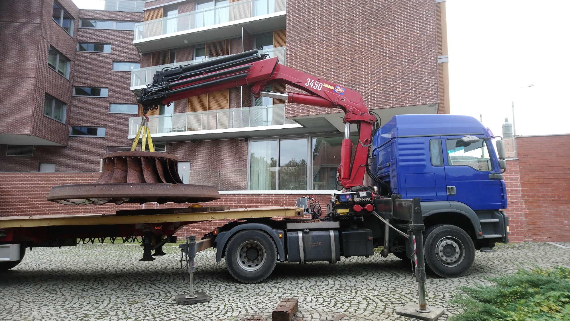 Niebieska ciężarówka z czerwonym dźwigiem podnosi dużą, zardzewiałą turbinę na przyczepę na tle ceglanego budynku.