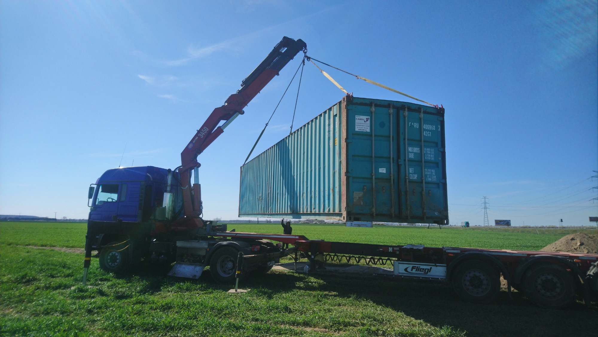 Ciężarówka z dźwigiem we Wrocławiu podnosi kontener na tle zielonego pola i błękitnego nieba.