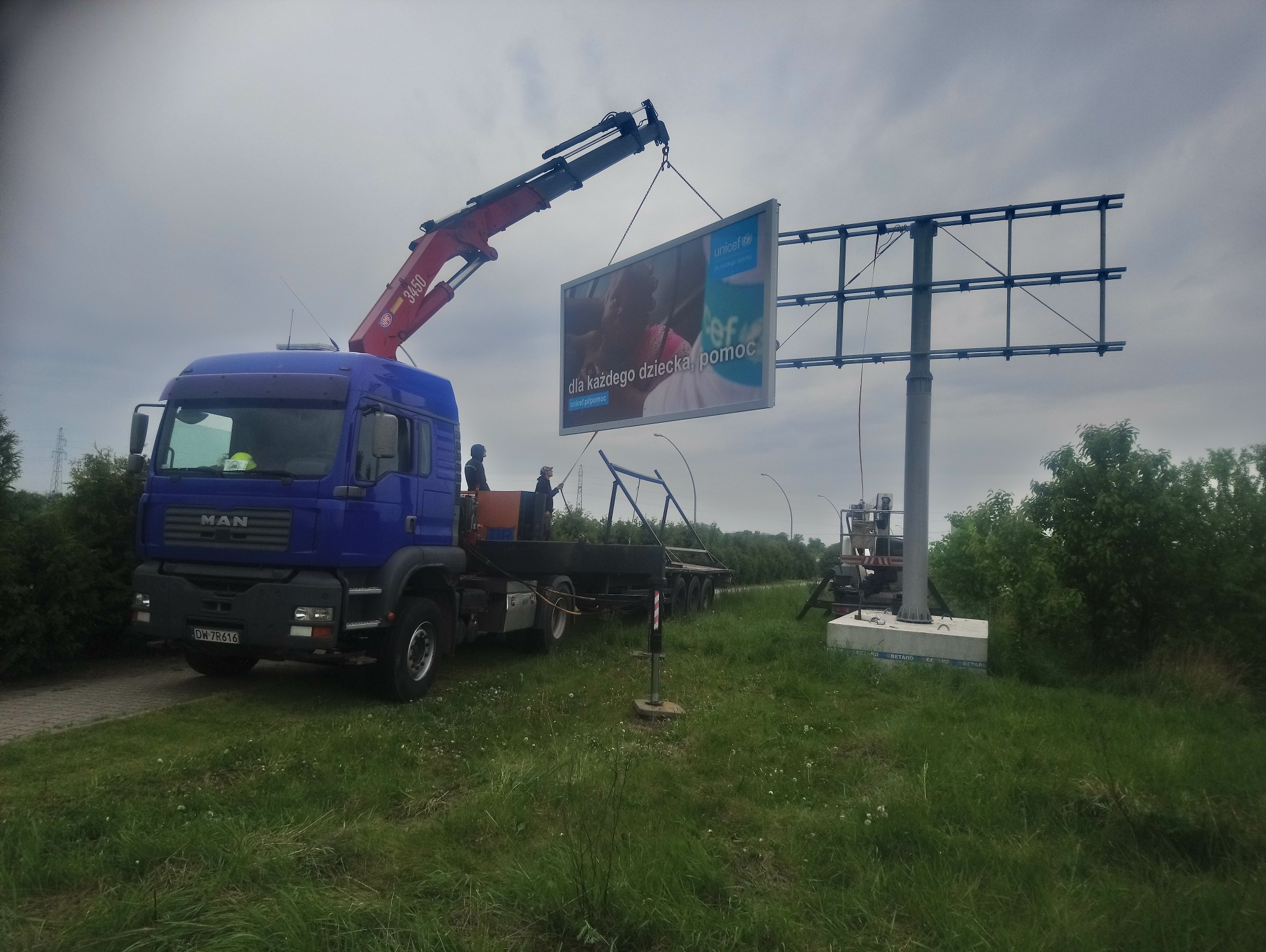 Montaż billboardu UNICEF z użyciem dźwigu samochodowego MAN na tle pochmurnego nieba.