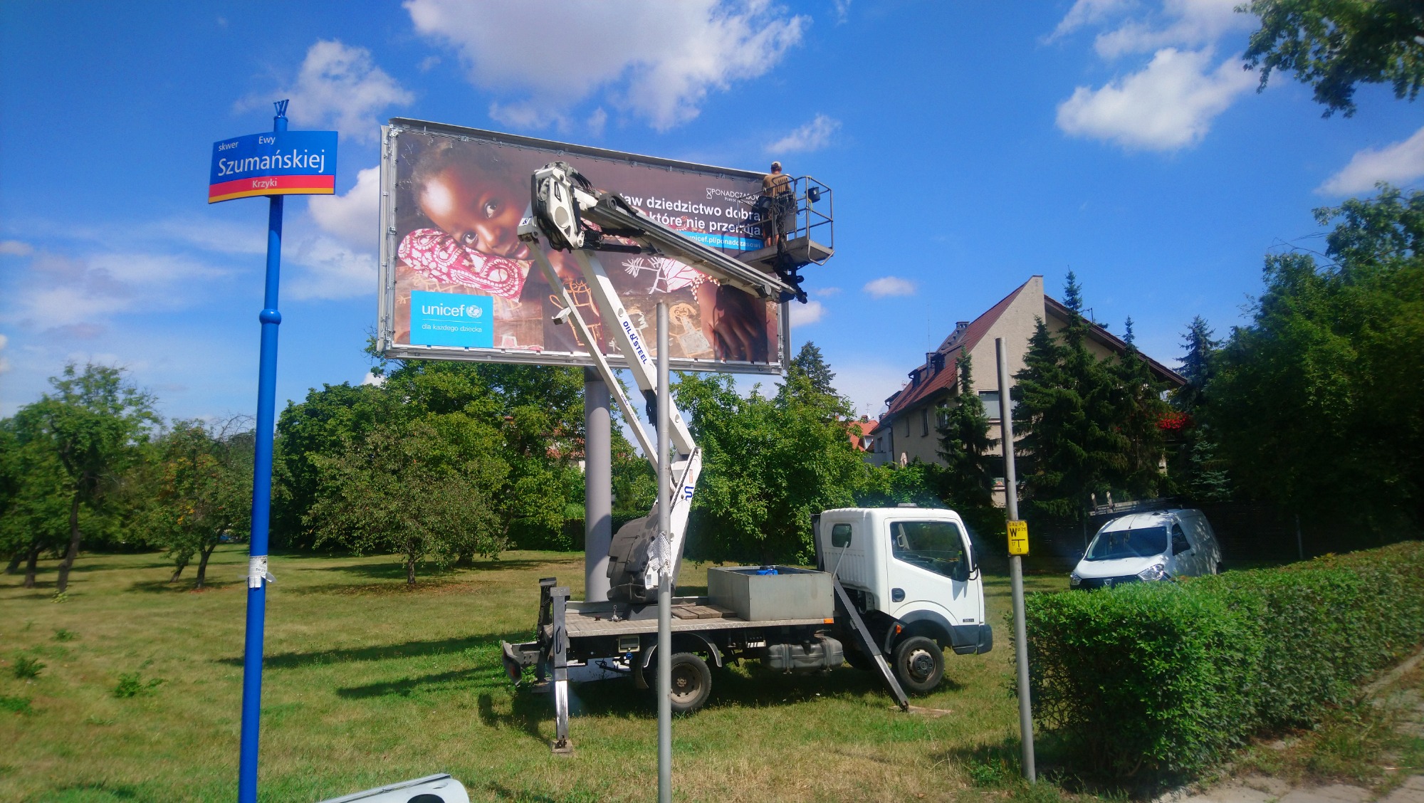 Montażysta na podnośniku koszowym instaluje plakat UNICEF na billboardzie przy skwerze Ewy Szumańskiej w Krzykach, na tle zieleni i błękitnego nieba.