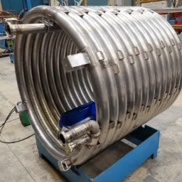 Duża, spiralna rura ze stali nierdzewnej na niebieskiej, drewnianej podstawie, z widocznymi spawami i numerami seryjnymi, w hali produkcyjnej.