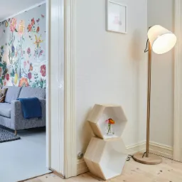 Jasne wnętrze mieszkania z szarą sofą, kwiatowym motywem na ścianie, drewnianą podłogą i lampą stojącą, widziane z perspektywy korytarza z białymi framugami drzwi.