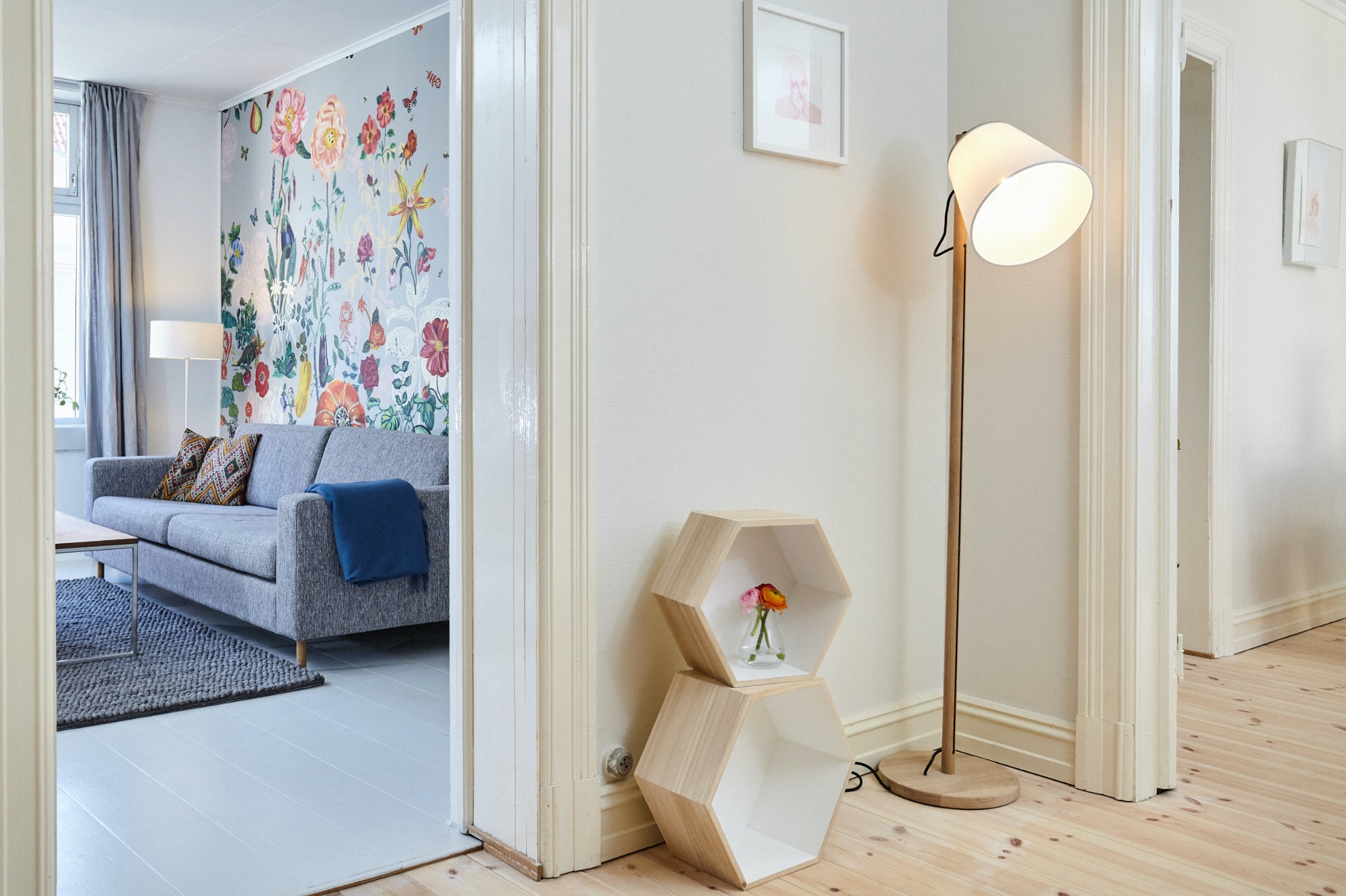 Jasne wnętrze mieszkania z szarą sofą, kwiatowym motywem na ścianie, drewnianą podłogą i lampą stojącą, widziane z perspektywy korytarza z białymi framugami drzwi.