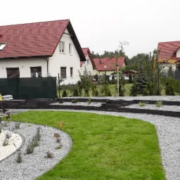 Nowoczesny ogród z trawnikiem, ścieżkami wysypanymi grysem i ozdobnymi nasadzeniami w otoczeniu domów jednorodzinnych.