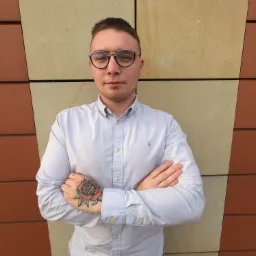 Portret młodego mężczyzny w okularach i koszuli z podwiniętymi rękawami, ze skrzyżowanymi rękami, na których widać tatuaż róży, stojącego na tle ściany z paneli w odcieniach beżu i brązu.