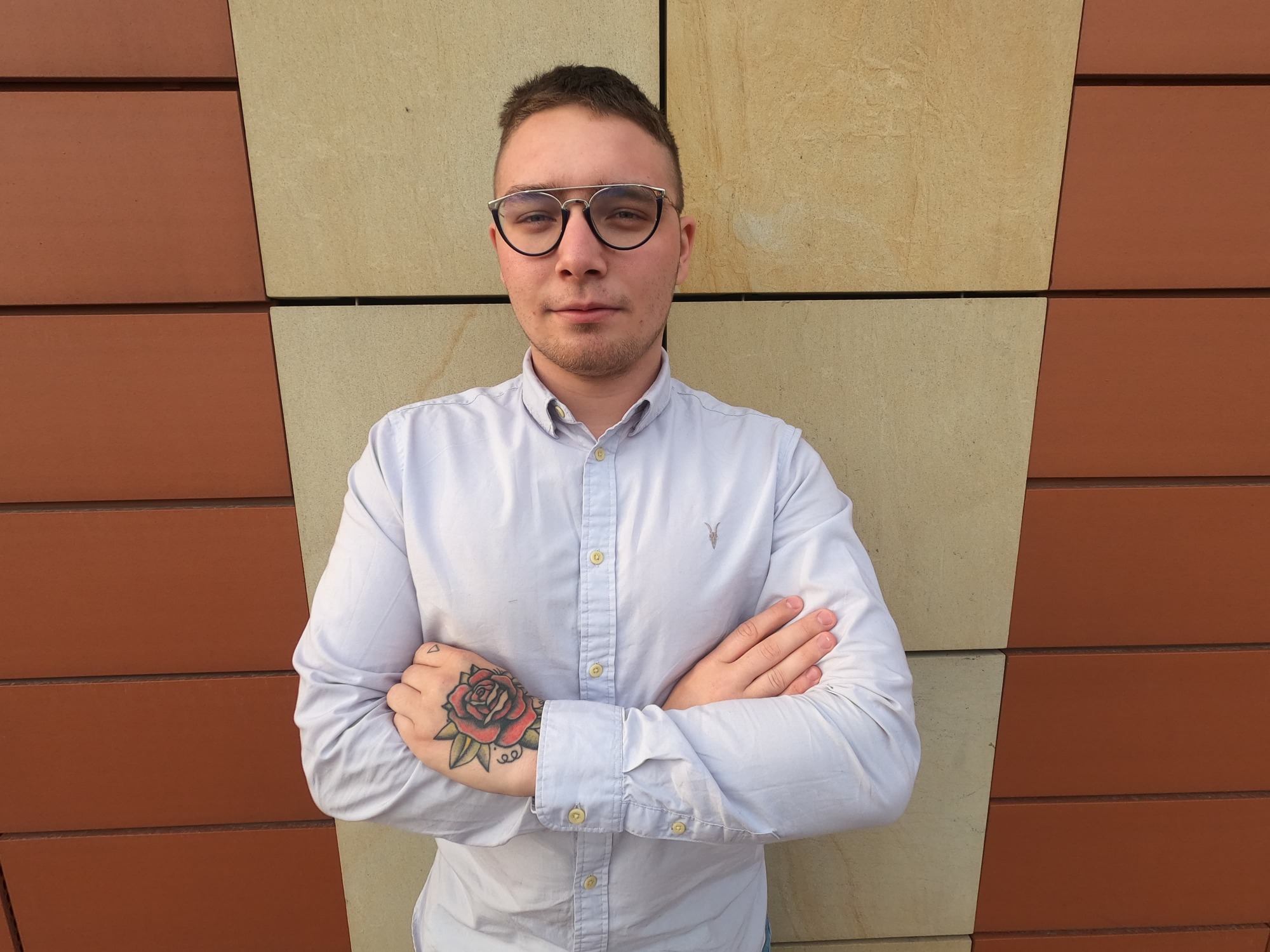 Portret młodego mężczyzny w okularach i koszuli z podwiniętymi rękawami, ze skrzyżowanymi rękami, na których widać tatuaż róży, stojącego na tle ściany z paneli w odcieniach beżu i brązu.